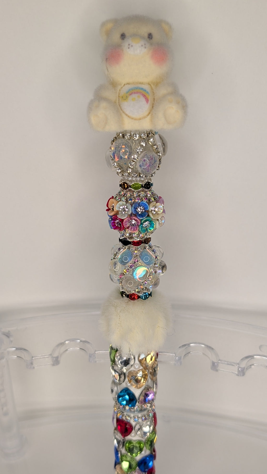 Decorative pen with colorful beads and a small plush bear on top, on a clear stand against a white background.