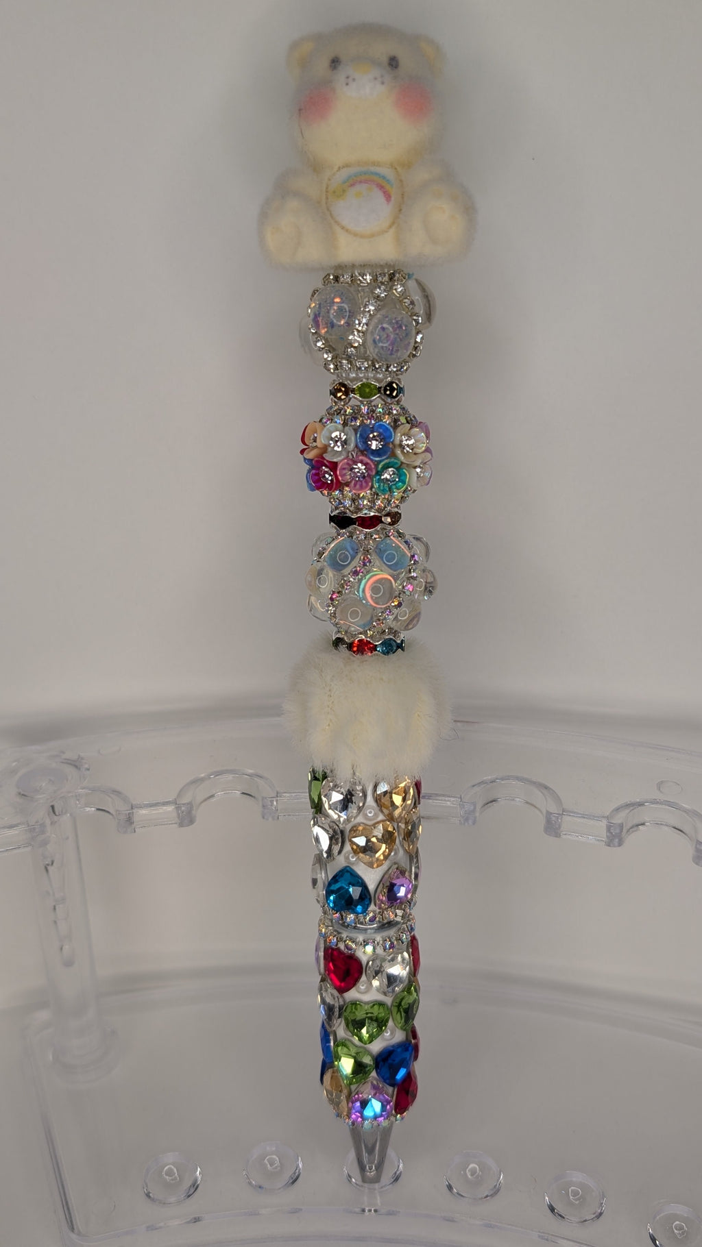 Decorative pen with colorful gemstones and a teddy bear at the top on a clear stand.