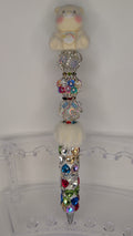 Decorative pen with colorful gemstones and a teddy bear at the top on a clear stand.