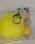 Yellow fluffy keychain with decorative beads on a clear stand.